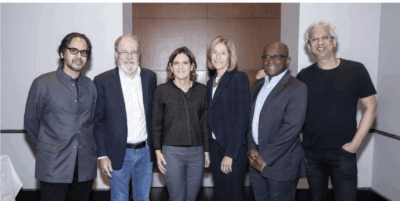 Six people stand side by side in a conference room, posing for a group photo. They are dressed in business or smart casual attire and are smiling at the camera. A table and a brown panel are visible behind them.