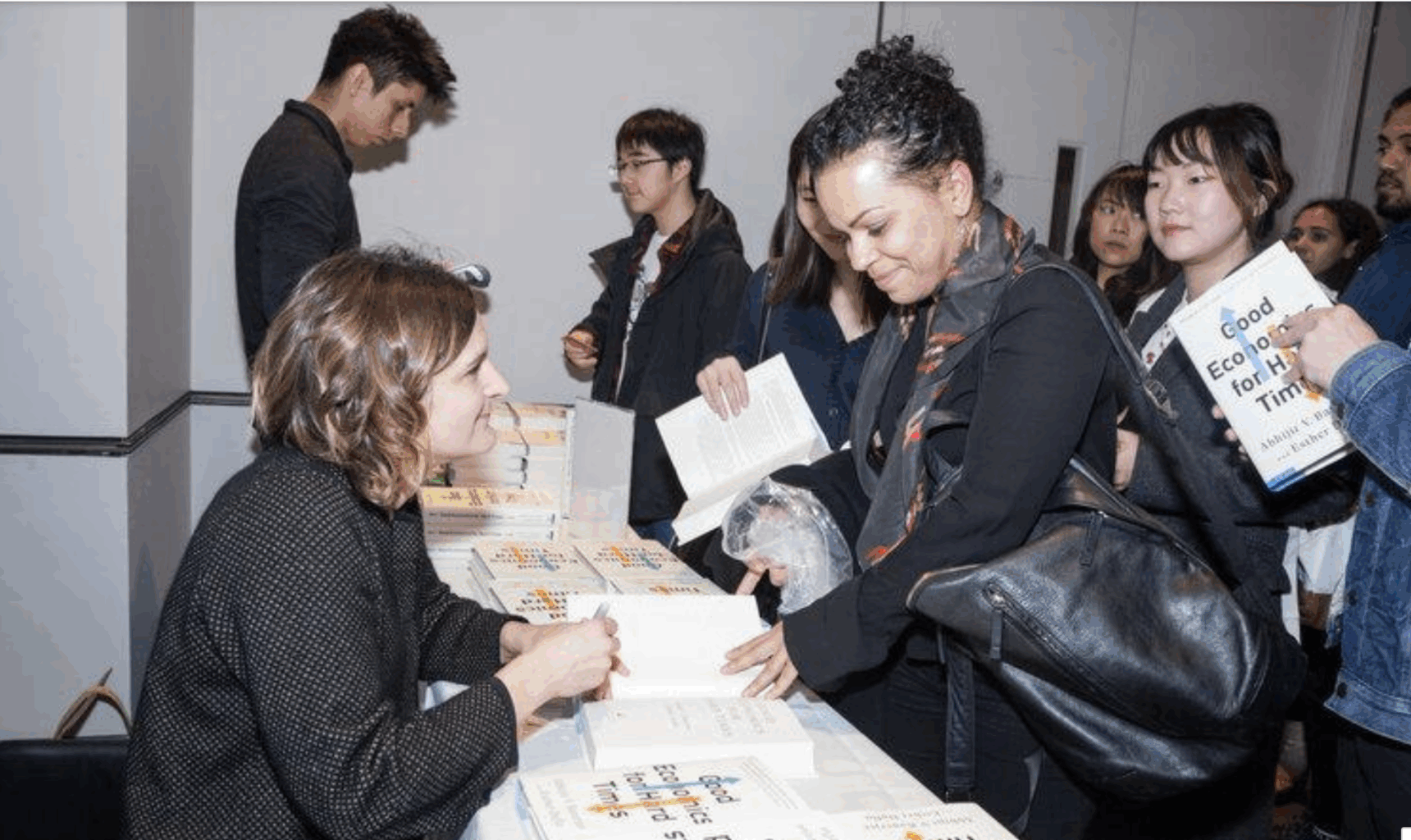 A woman sits at a table signing books for people standing in line at an indoor event. Attendees hold books and chat with her, and several are holding bags or backpacks. Laptops and paperwork are visible on the table.