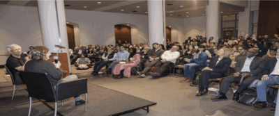 A woman speaks at a podium to a seated audience in a large, modern lecture hall; a man sits beside her on stage while attendees listen attentively. A laptop screen with editing tools frames the image.