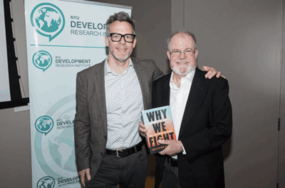 Two men stand and smile at the camera; one holds a book titled Why We Fight. Behind them is a banner with the NYU Development Research Institute logo. A laptop screen with various application tabs is visible.