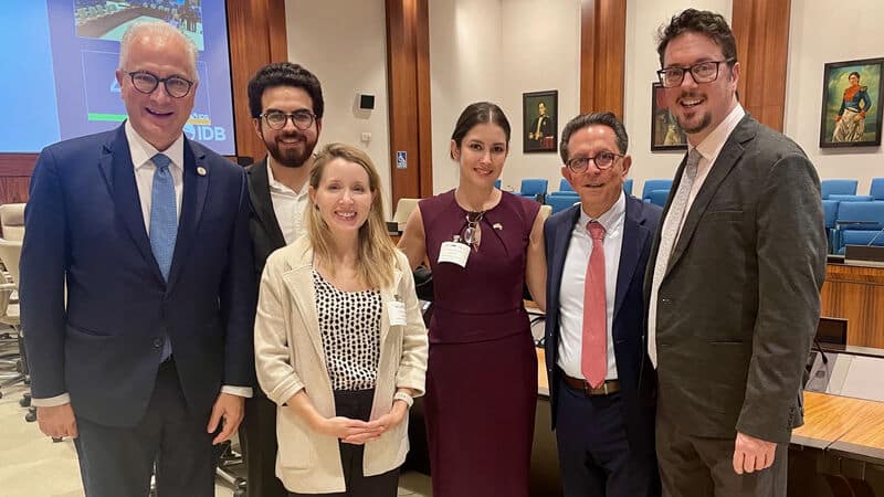 Six people in business attire stand together and smile for a group photo in a conference room with wooden panels, blue chairs, and framed portraits on the walls.