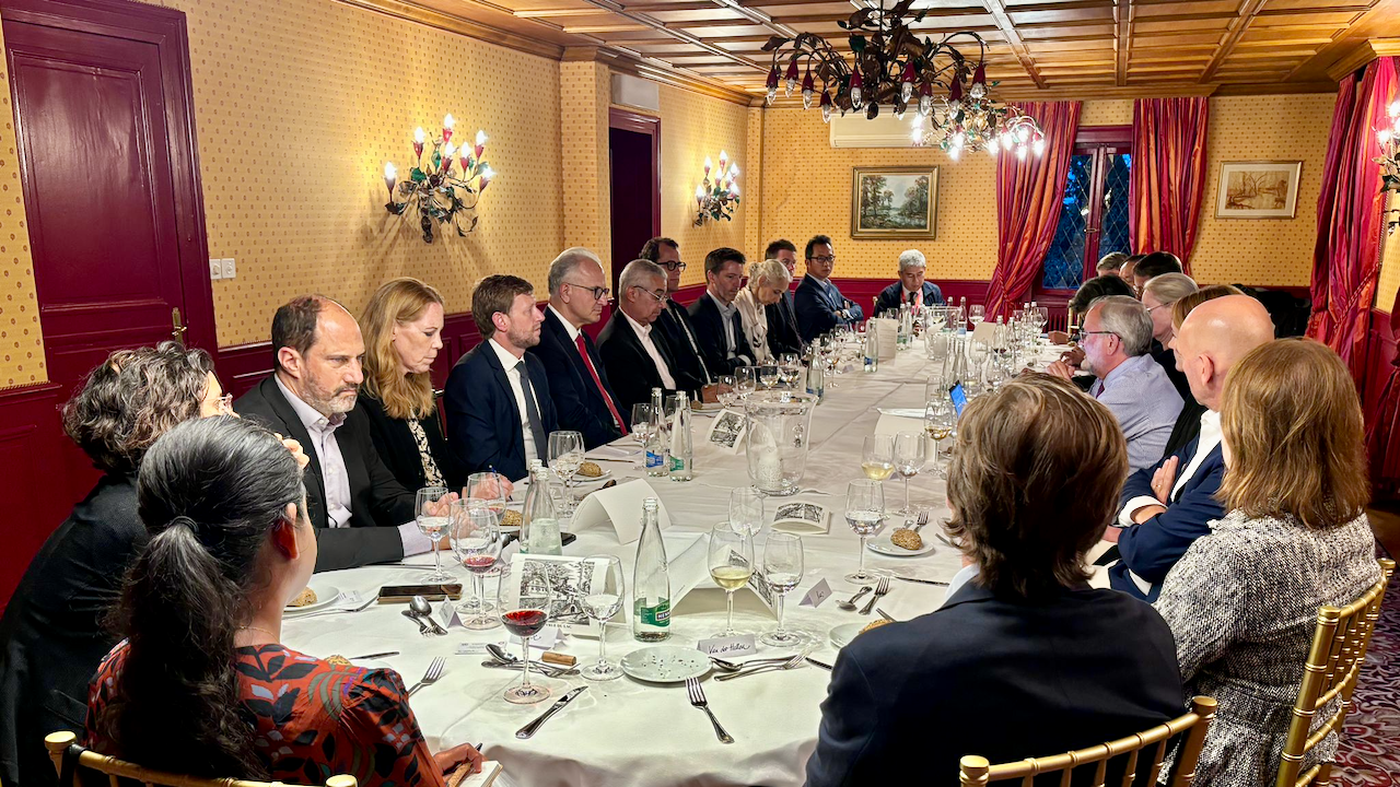 A group of people in formal attire sit around a long, elegantly set dining table in a warmly lit, classic room with red accents, chandeliers, and patterned wallpaper, engaged in conversation.