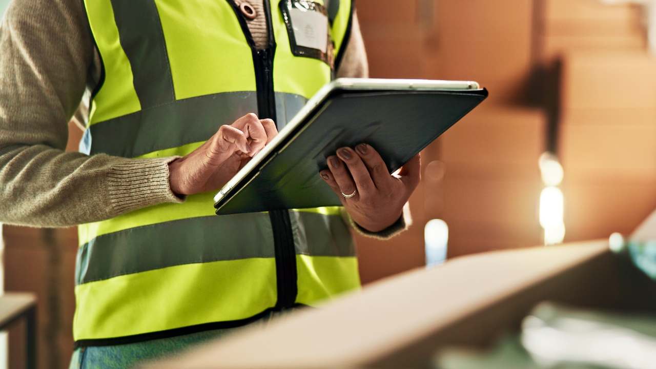 A person wearing a reflective safety vest uses a tablet device, appearing to check or record information in an indoor work environment. The focus is on the hands and device, not the face.