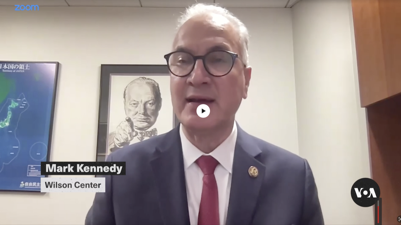 A man in a suit and red tie speaks during a video call. On the screen, Mark Kennedy, Wilson Center is labeled. Behind him is a portrait of Winston Churchill. The VOA and Zoom logos appear on the image.