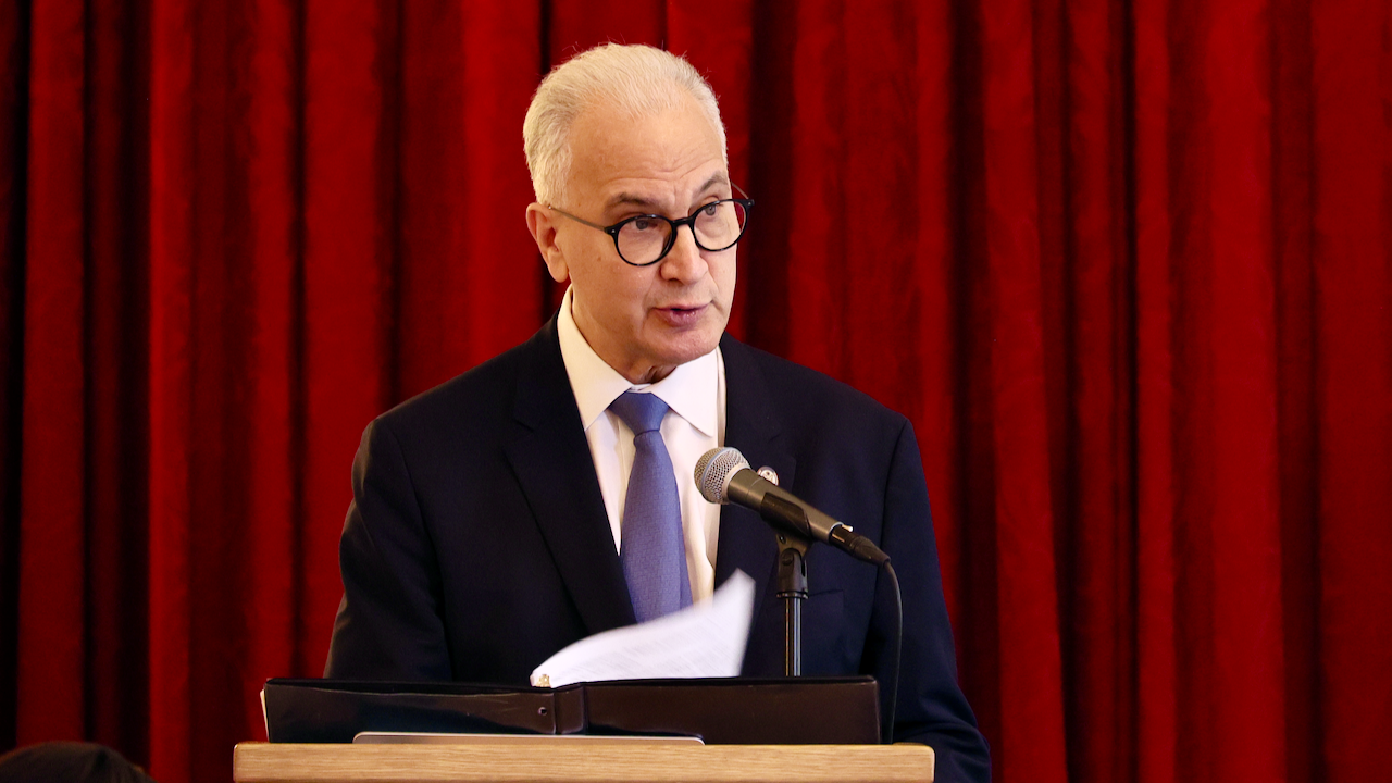 An older man in a suit, tie, and glasses speaks at a podium with a microphone, holding papers, against a backdrop of red curtains.