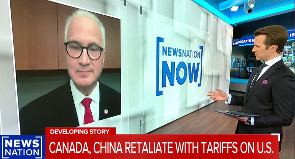 A news anchor stands in a studio holding papers, interviewing a man via video call on a large screen. A headline reads, CANADA, CHINA RETALIATE WITH TARIFFS ON U.S. and the show is titled NewsNation Now.