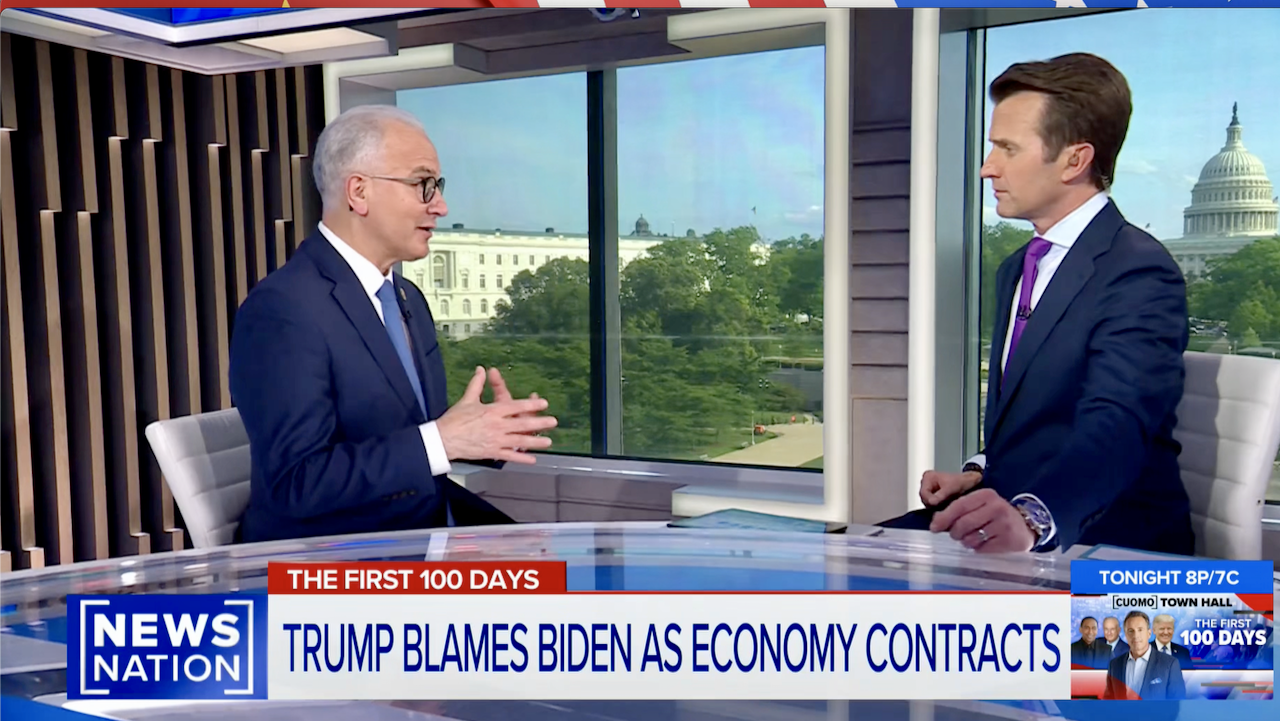 Two news anchors sit at a desk in a bright studio with large windows overlooking the White House; the headline on screen reads, Trump blames Biden as economy contracts.