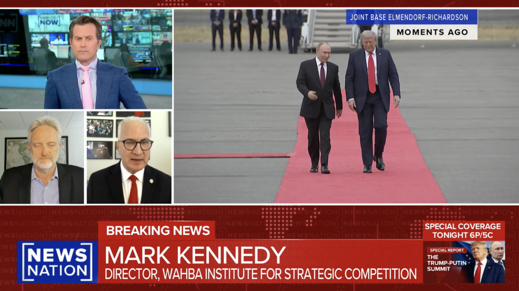 A news broadcast shows four commentators discussing breaking news, with an image of two men walking on a red carpet at Joint Base Elmendorf-Richardson displayed. The ticker reads: Mark Kennedy, Director, Wahiba Institute for Strategic Competition.