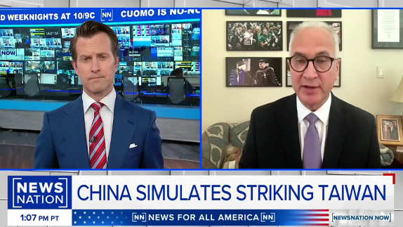 A news broadcast split-screen shows a male anchor on the left in a studio and a male guest in glasses and suit on the right in a home office. The headline reads, “China Simulates Striking Taiwan.”.