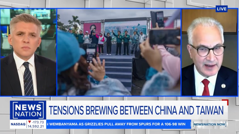A news broadcast shows a split screen: a serious male anchor on the left, a male guest in glasses on the right, and a central image of people at an outdoor event taking photos. Text reads TENSIONS BREWING BETWEEN CHINA AND TAIWAN.