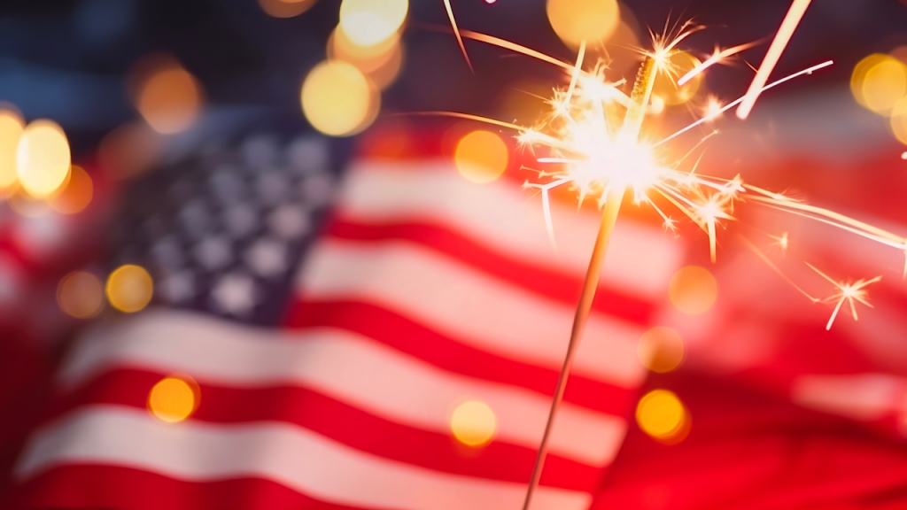A sparkler glows brightly in the foreground with golden light, while a blurred American flag is visible in the background, creating a festive and patriotic atmosphere.