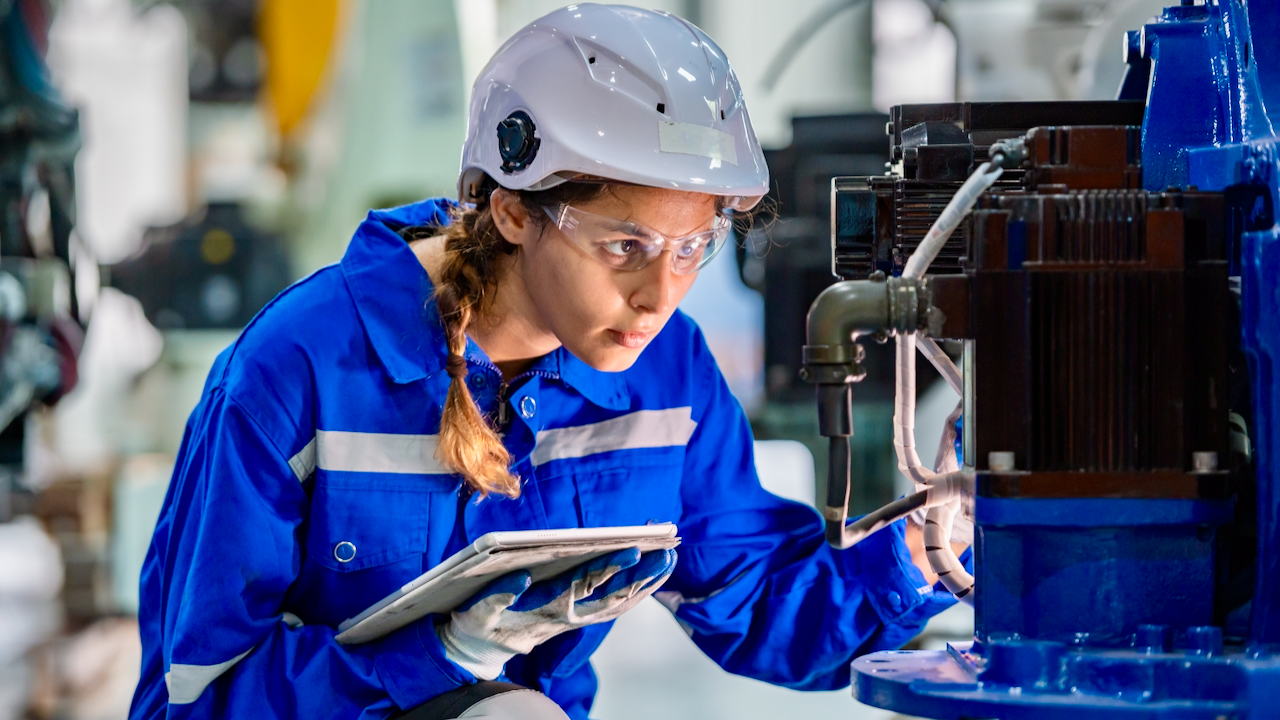 A woman in blue coveralls, a white helmet, and safety glasses inspects industrial machinery while holding a tablet, appearing focused and attentive in a factory or industrial setting.
