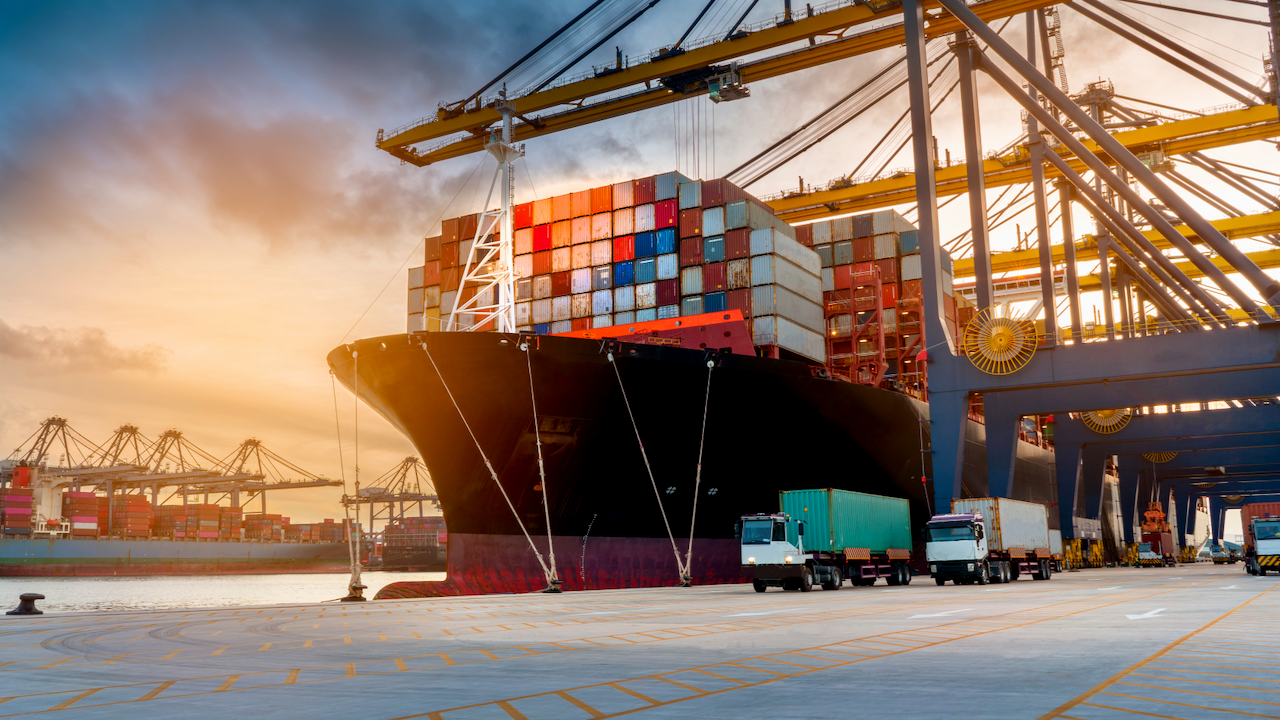 A large cargo ship loaded with colorful shipping containers is docked at a port, with cranes unloading containers onto trucks at sunset.