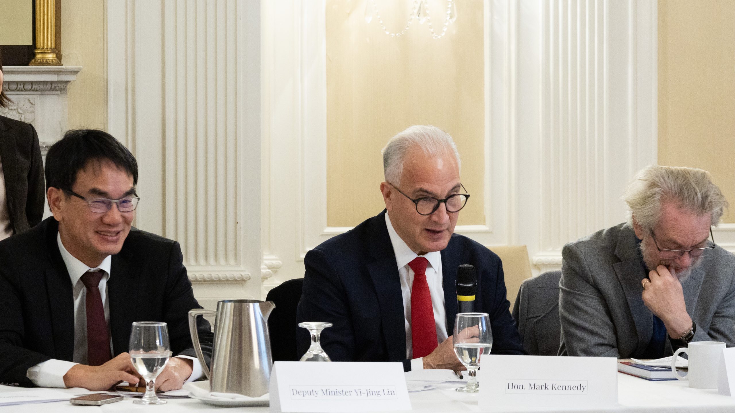 Three men in suits sit at a conference table with microphones, glasses of water, and nameplates. The man in the center speaks, holding a microphone, while the others listen. The room has white walls and ornate trim.