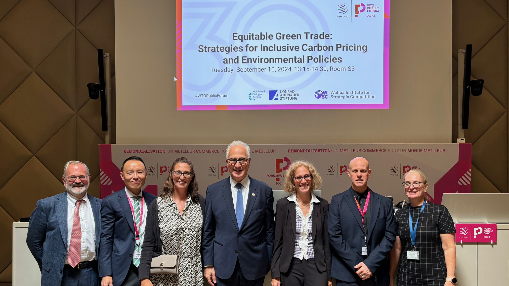 Eight people stand smiling in front of a presentation screen titled “Equitable Green Trade: Strategies for Inclusive Carbon Pricing and Environmental Policies” at a conference. A pink and white event backdrop is behind them.