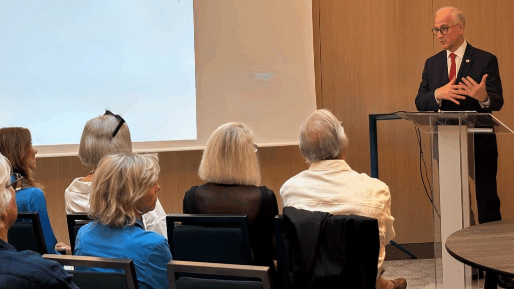 A man in a suit and red tie speaks at a podium to an audience seated in chairs, with a white screen and wooden walls in the background.