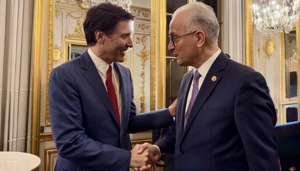 Two men in suits smiling and shaking hands in an ornate, gold-decorated room with mirrors and chandeliers. One has dark hair and a red tie; the other is bald and wears glasses with a purple tie.
