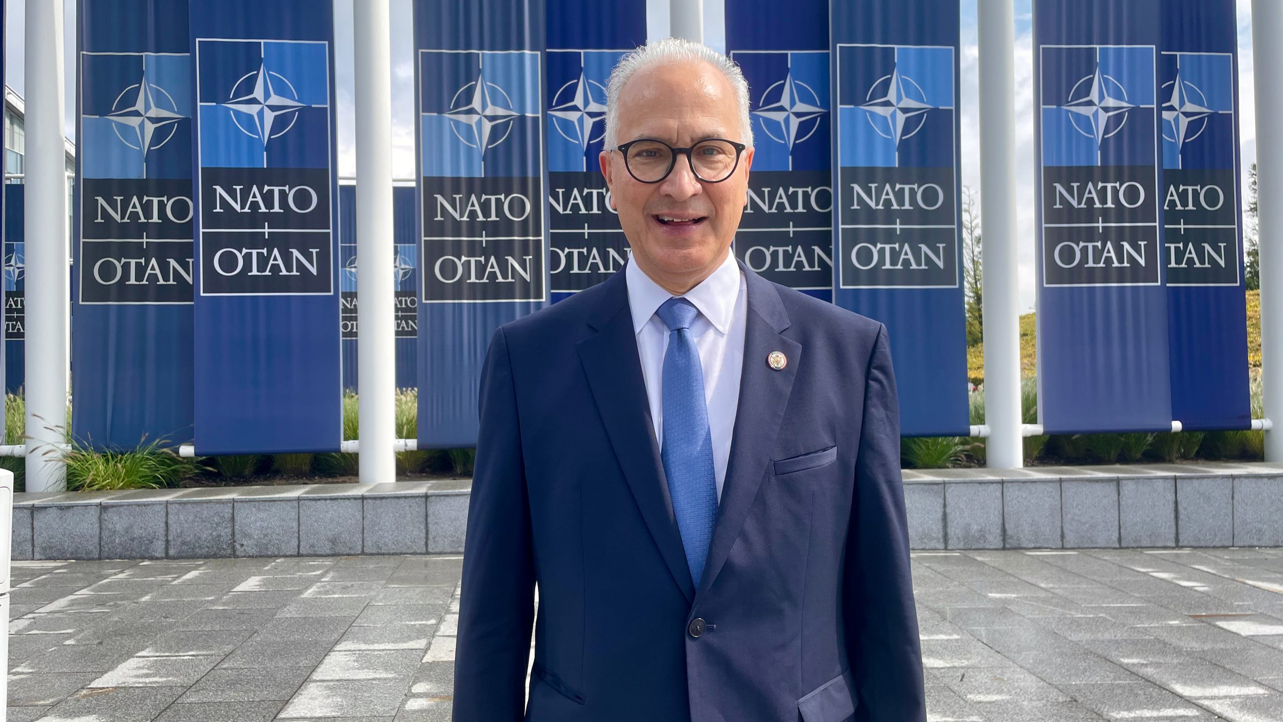 A man in a navy suit and light blue tie stands smiling in front of large NATO/OTAN banners and flagpoles outside a modern building.