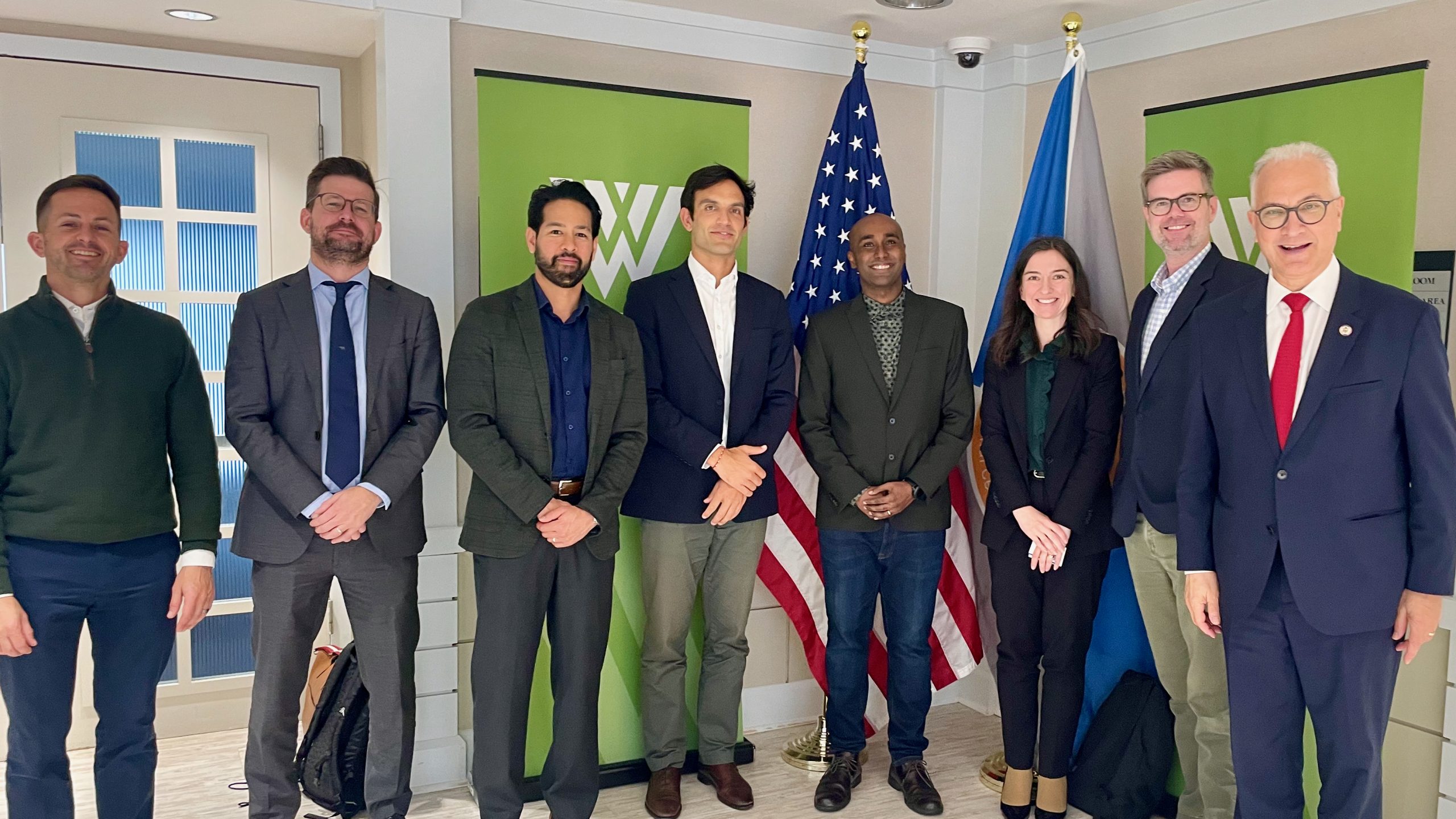 Seven people pose indoors in business attire, standing in front of green banners, an American flag, and another blue flag. They are smiling, and the setting appears to be a professional or formal event.