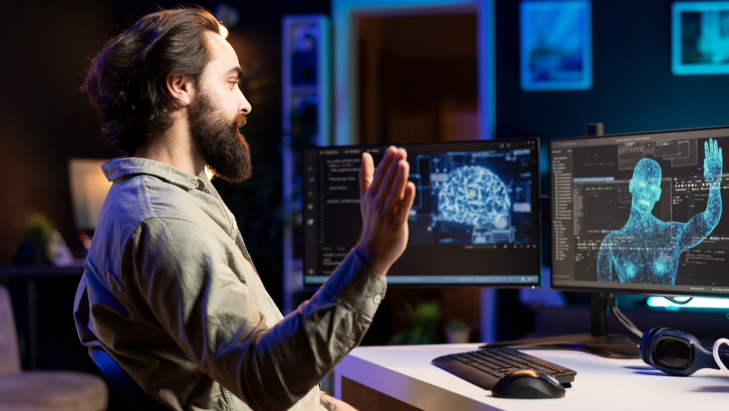 A man sits at a desk waving at a monitor, where a digital human figure on the screen appears to wave back. Computer code and a brain illustration are visible on dual monitors in a modern, dimly lit room.