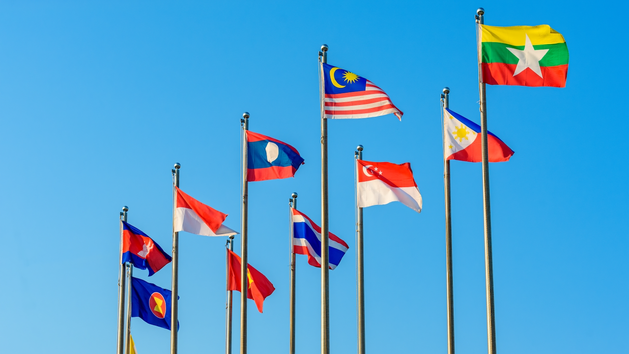 Ten national flags and one organization flag of Southeast Asian countries, including Malaysia, Myanmar, Thailand, Vietnam, and Singapore, are displayed on flagpoles against a clear blue sky.