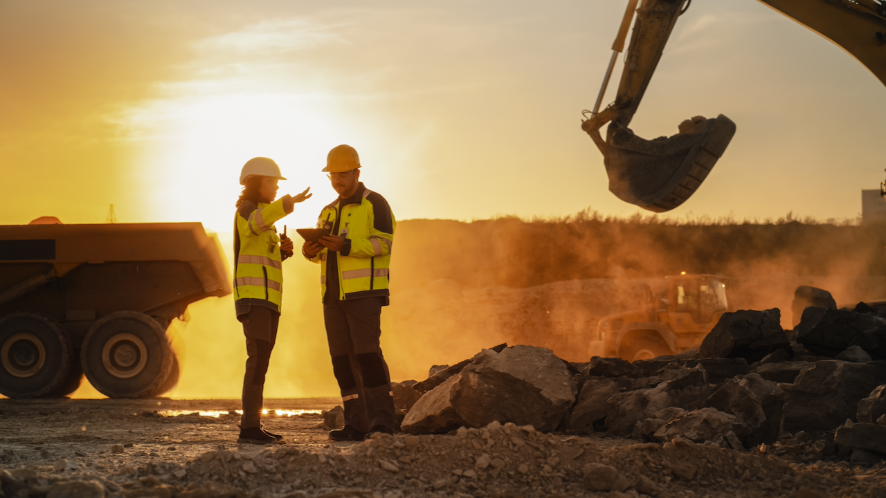 Two construction workers wearing safety gear discuss plans at a dusty construction site during sunset, with heavy machinery and large rocks in the background.