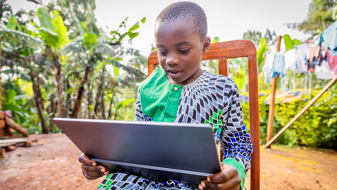 A young boy sitting outdoors on a wooden chair, looking at a laptop. He wears a colorful patterned shirt, with plants, trees, and laundry hanging in the background, suggesting a rural setting.