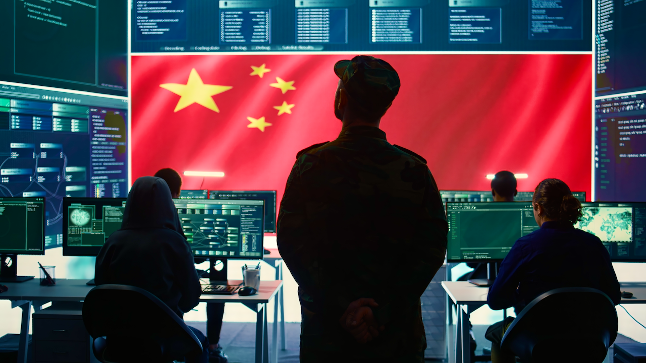 A person in military uniform stands in front of computer workstations and large monitors, with a Chinese flag displayed prominently on a screen in the background. Other individuals work at the computers.