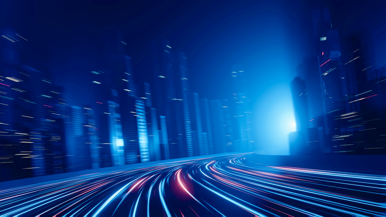 Futuristic cityscape at night with tall skyscrapers and glowing light trails on a curving road, suggesting high-speed motion and advanced technology. The scene is dominated by blue tones and dynamic lighting effects.