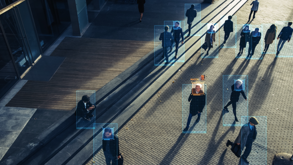A city street with multiple people walking, each person highlighted by blue digital rectangles, some showing facial recognition tags, representing surveillance and identification technology.