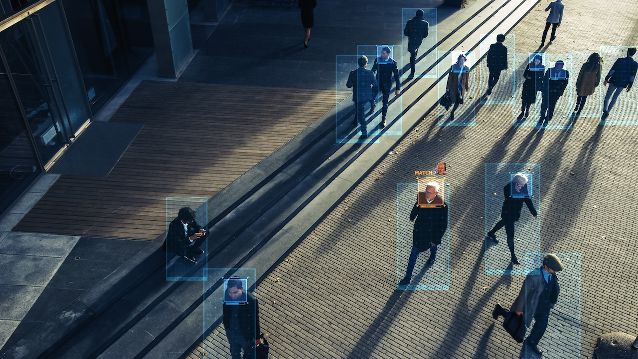 A city street with multiple people walking, each person highlighted by blue digital rectangles, some showing facial recognition tags, representing surveillance and identification technology.