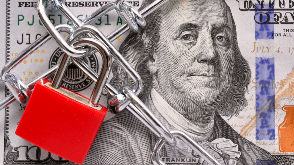 A close-up of a U.S. $100 bill covered with a metal chain and secured by a red padlock, symbolizing restricted access or financial security.