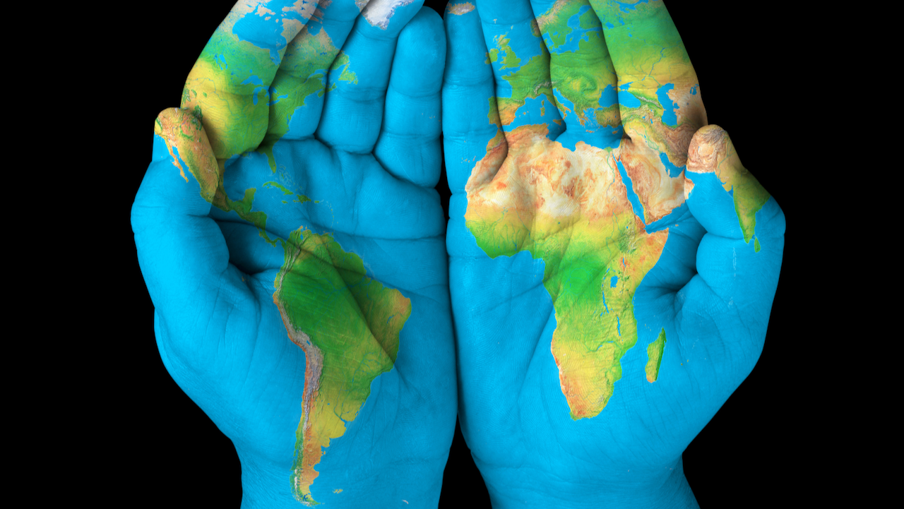 Two hands are cupped together against a black background, with a detailed map of the world painted on them, showing continents and oceans across the palms and fingers.