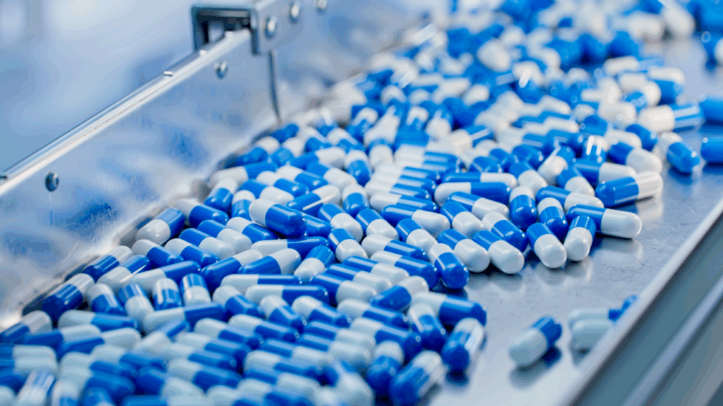 Hundreds of blue and white capsules move along a stainless steel conveyor belt in a pharmaceutical manufacturing facility.