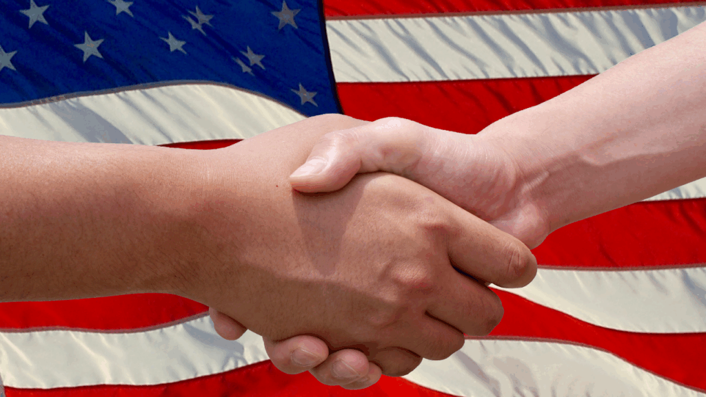Two people shaking hands in front of a large American flag, symbolizing agreement, partnership, or unity in the context of the United States.