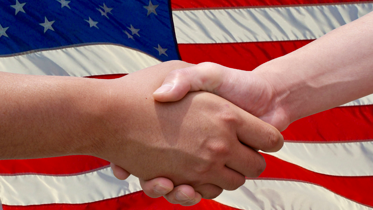 Two people shaking hands in front of a large American flag, symbolizing agreement, partnership, or unity in the context of the United States.