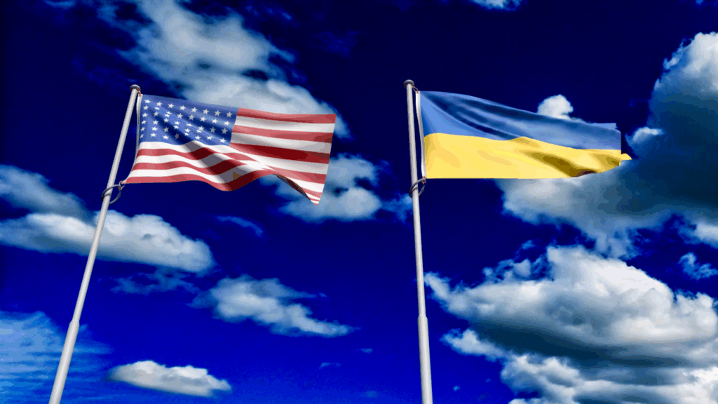 The American and Ukrainian flags wave on flagpoles against a bright blue sky with scattered white clouds.