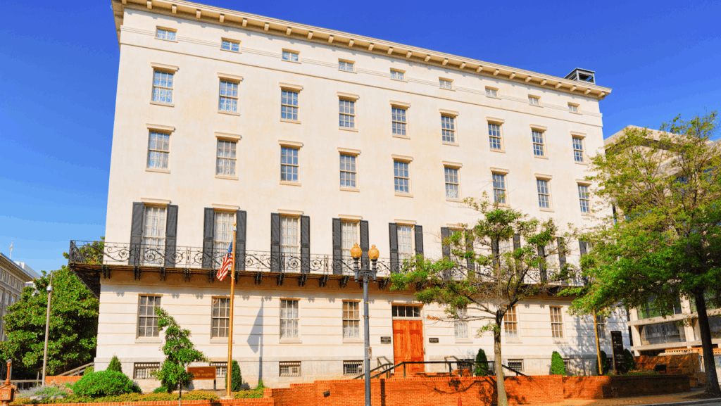 Four-story cream-colored historic building with tall windows, decorative ironwork balcony, and a wooden front door. American flag and black flags hang in front. Trees and a brick sidewalk are visible, under a clear blue sky.