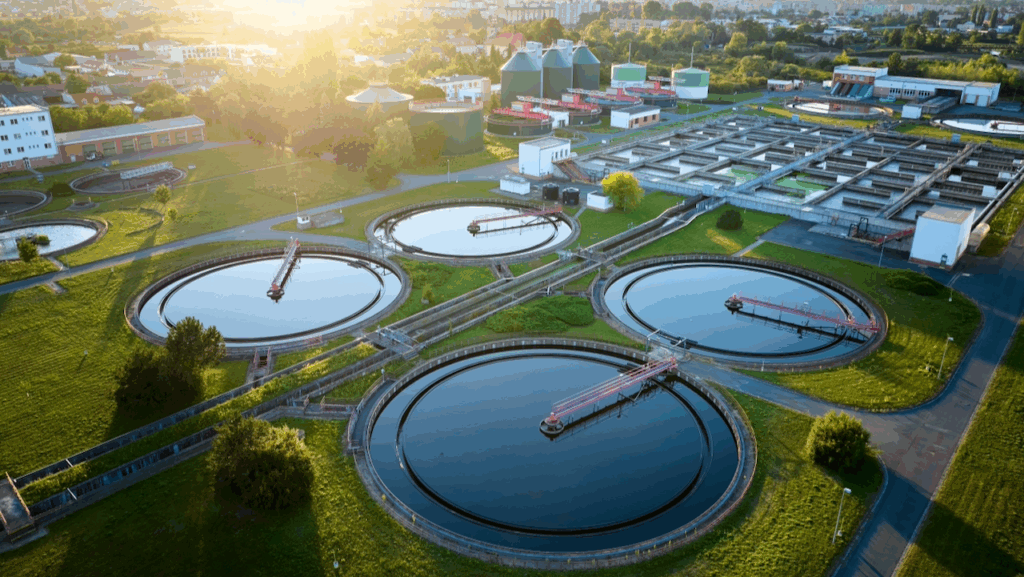 Aerial view of a modern wastewater treatment plant with large circular and rectangular tanks, surrounded by green grass and buildings, under bright sunlight.