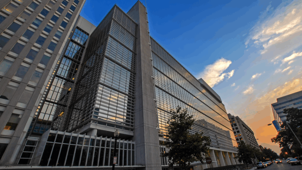 A modern glass and steel office building stands tall under a blue and orange sky at sunset, with surrounding trees and adjacent buildings visible along the street.