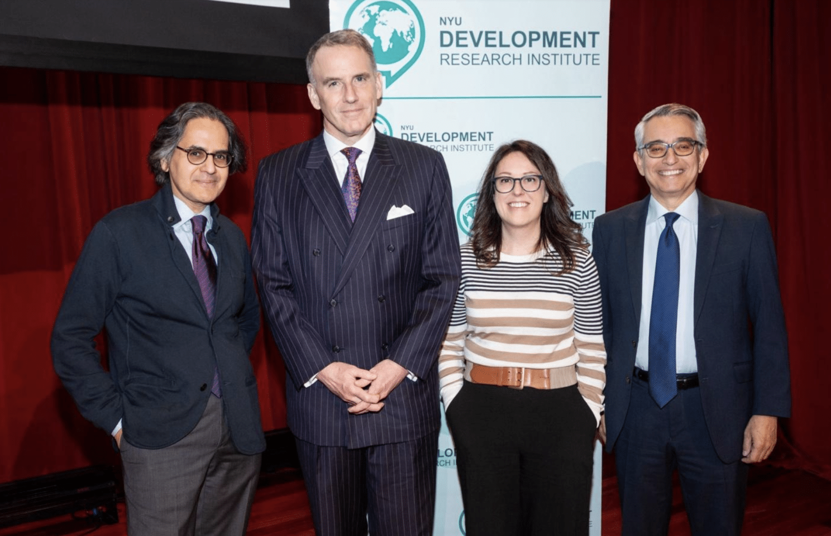 Four people stand smiling in front of an NYU Development Research Institute banner and a red curtain. Three men wear suits and ties, and a woman wears glasses, a striped sweater, and dark pants.