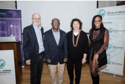 Four people stand side by side, smiling for a group photo at an event. Two roll-up banners with logos and text are displayed behind them. The setting appears to be a conference or seminar room.