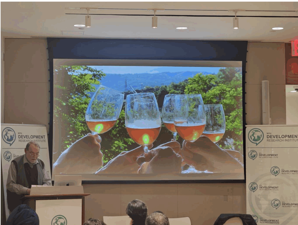 A speaker stands at a podium in front of an audience, with a large screen behind him displaying an image of four hands holding up glasses of wine against a scenic outdoor background.