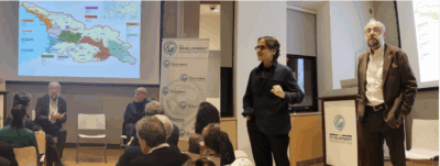 A panel discussion is taking place in a conference room. Two men are standing and speaking at the front, while others are seated. A map is projected on a screen behind them, and several attendees are watching and listening.