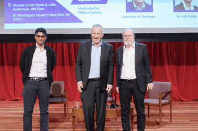 Three men stand on a stage in front of red curtains and a projected screen displaying event information. Two chairs and a small table are behind them. Each man is dressed in business or business casual attire.