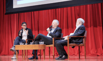 Three men sit on a stage in front of red curtains, engaged in a panel discussion. Two hold microphones, and all are seated in wooden chairs around a small table. A projected screen is partly visible above them.