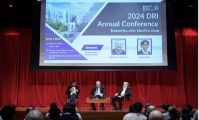 Three people sit on stage in front of a red curtain, speaking at the 2024 DRI Annual Conference. A large screen behind them displays conference details and speaker photos, while the audience watches from the foreground.