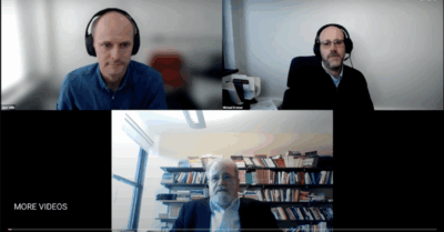 A Zoom webinar is displayed on a computer screen, showing three men with headsets in separate video frames. The bottom participant sits in front of bookshelves, while the others are in office settings. Various apps are open on the desktop.