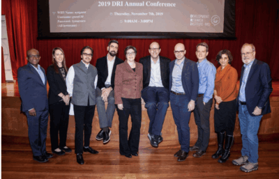 A group of ten people stand on a wooden stage in front of a screen displaying 2019 DRI Annual Conference. They are posing together and smiling, dressed in business or business casual attire.