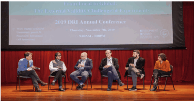 Six people sit on a stage panel discussion in front of a screen displaying 2019 DRI Annual Conference details. They hold microphones and converse, with a red curtain backdrop and an audience visible in the foreground.